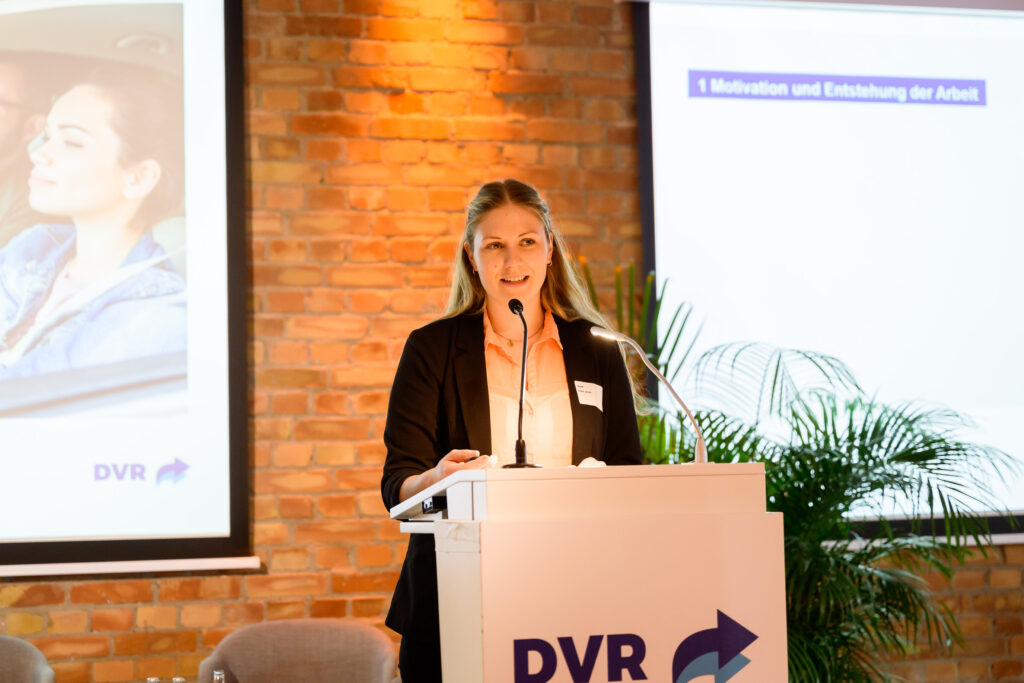 Frau steht an einem Rednerpult und hält einen Vortrag vor einer Präsentationsfolie in einem Veranstaltungsraum.