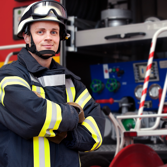 Feuerwehrmann in Schutzkleidung und Helm steht mit verschränkten Armen vor einem Einsatzfahrzeug. Im Hintergrund sind Ausrüstungsgegenstände zu sehen. Die Szene vermittelt Einsatzbereitschaft und Professionalität.