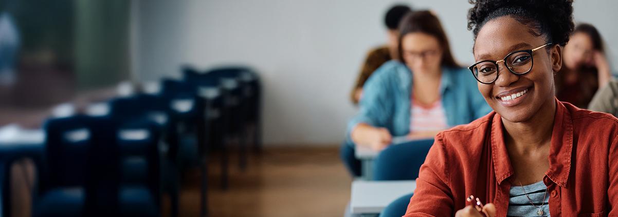 Studentin sitzt in einem Seminarraum und lächelt in die Kamera. Im Hintergrund sind weitere Studierende an Tischen zu sehen, die an ihren Aufgaben arbeiten. Die Szene vermittelt Studium, Lernen und eine positive Lernatmosphäre.
