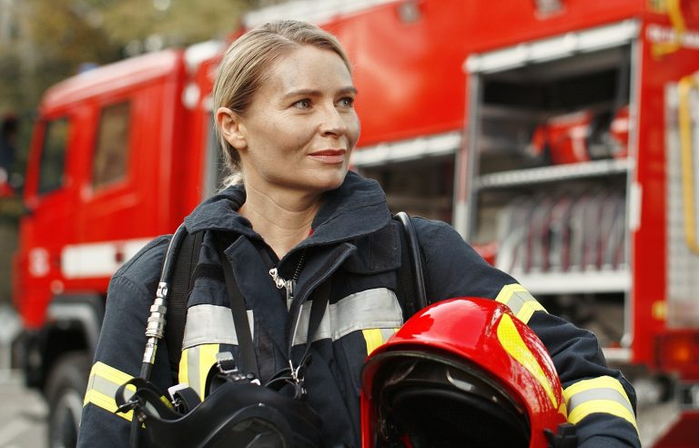 Eine Feuerwehrfrau steht in Einsatzkleidung vor einem roten Feuerwehrfahrzeug. Sie hält einen Feuerwehrhelm und weitere Ausrüstung in den Armen und blickt aufmerksam zur Seite. Im Hintergrund ist das geöffnete Fahrzeug mit Einsatzmaterial zu sehen. Die Szene vermittelt Einsatzbereitschaft und Professionalität im Bevölkerungsschutz.