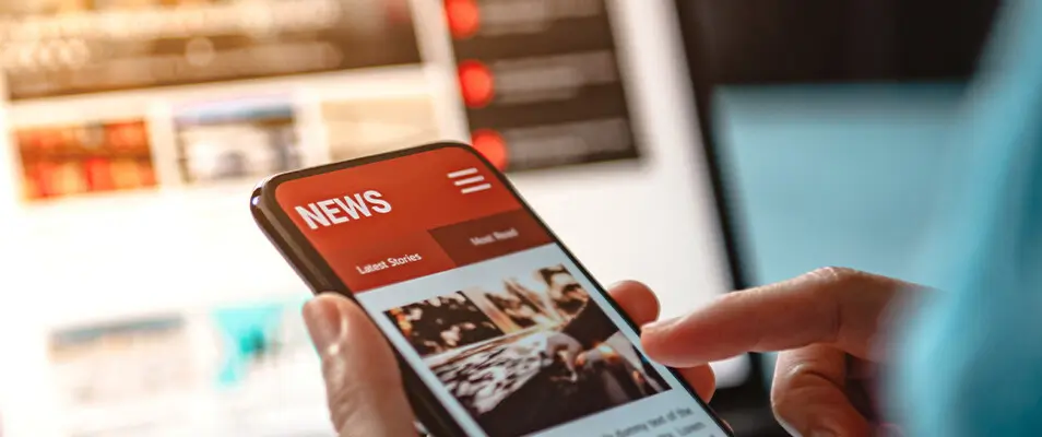 Person hält ein Smartphone mit geöffneter App in der Hand, im Hintergrund ein Laptop.