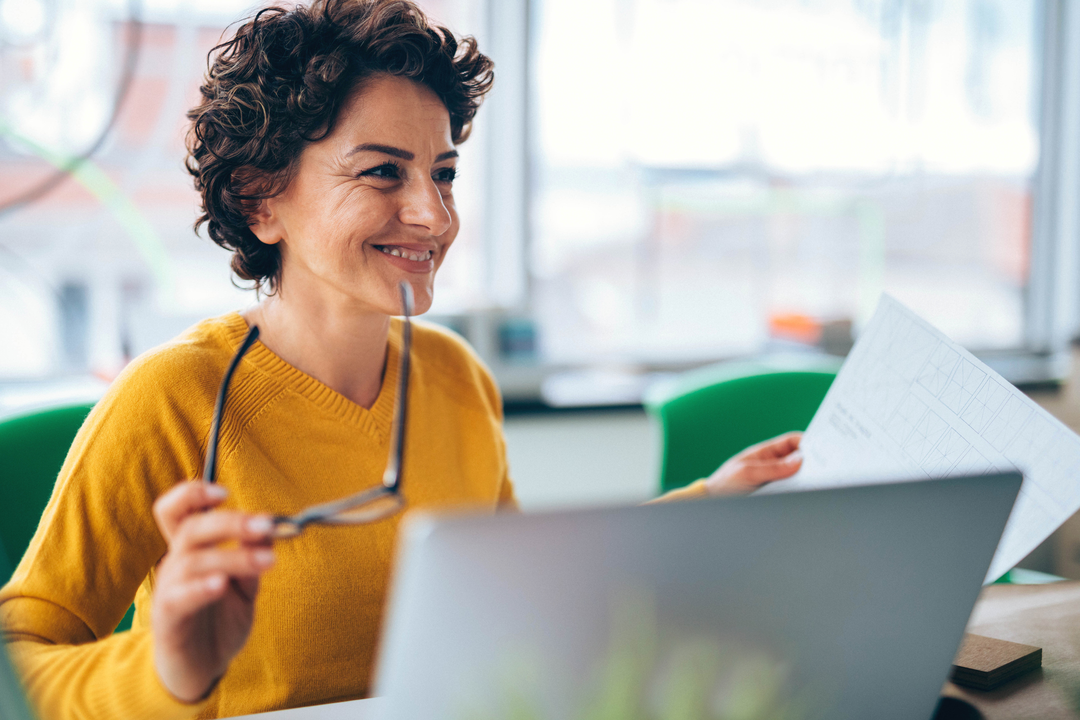 Eine Frau sitzt an einem Schreibtisch und arbeitet an einem Laptop. Sie lächelt und hält eine Brille in der Hand sowie ein Blatt Papier mit Notizen. Der Arbeitsplatz ist hell und modern gestaltet, im Hintergrund sind Fenster zu sehen.