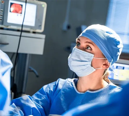 Eine Pflegefachperson in Schutzkleidung, OP-Haube und Mundschutz arbeitet konzentriert im Operationssaal. Im Hintergrund sind medizinische Geräte und ein Monitor zu sehen. Die Person blickt aufmerksam nach oben, während sie Teil eines chirurgischen Eingriffs ist.