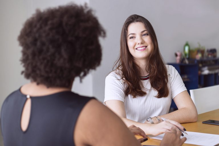 Zwei Frauen sitzen an einem Tisch und führen ein Gespräch. Eine Frau lächelt ihre Gesprächspartnerin an, während Unterlagen auf dem Tisch liegen.