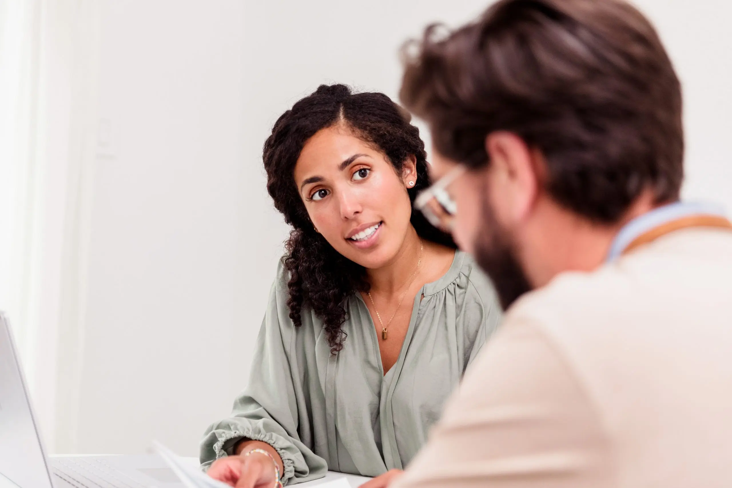 Zwei Erwachsene sitzen an einem Tisch und führen ein Gespräch. Eine Frau blickt aufmerksam und zugewandt zu einem Mann, der ihr gegenüber sitzt und Unterlagen vor sich hat. Die Frau gestikuliert leicht mit der Hand, was auf einen erklärenden oder beratenden Austausch hinweist. Die Szene wirkt ruhig und konzentriert und findet in einem hellen, neutralen Raum statt.