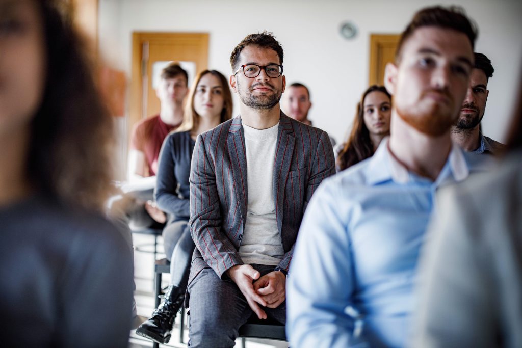 Studierende sitzen aufmerksam in einem Seminarraum und hören einer Person im Vordergrund zu.