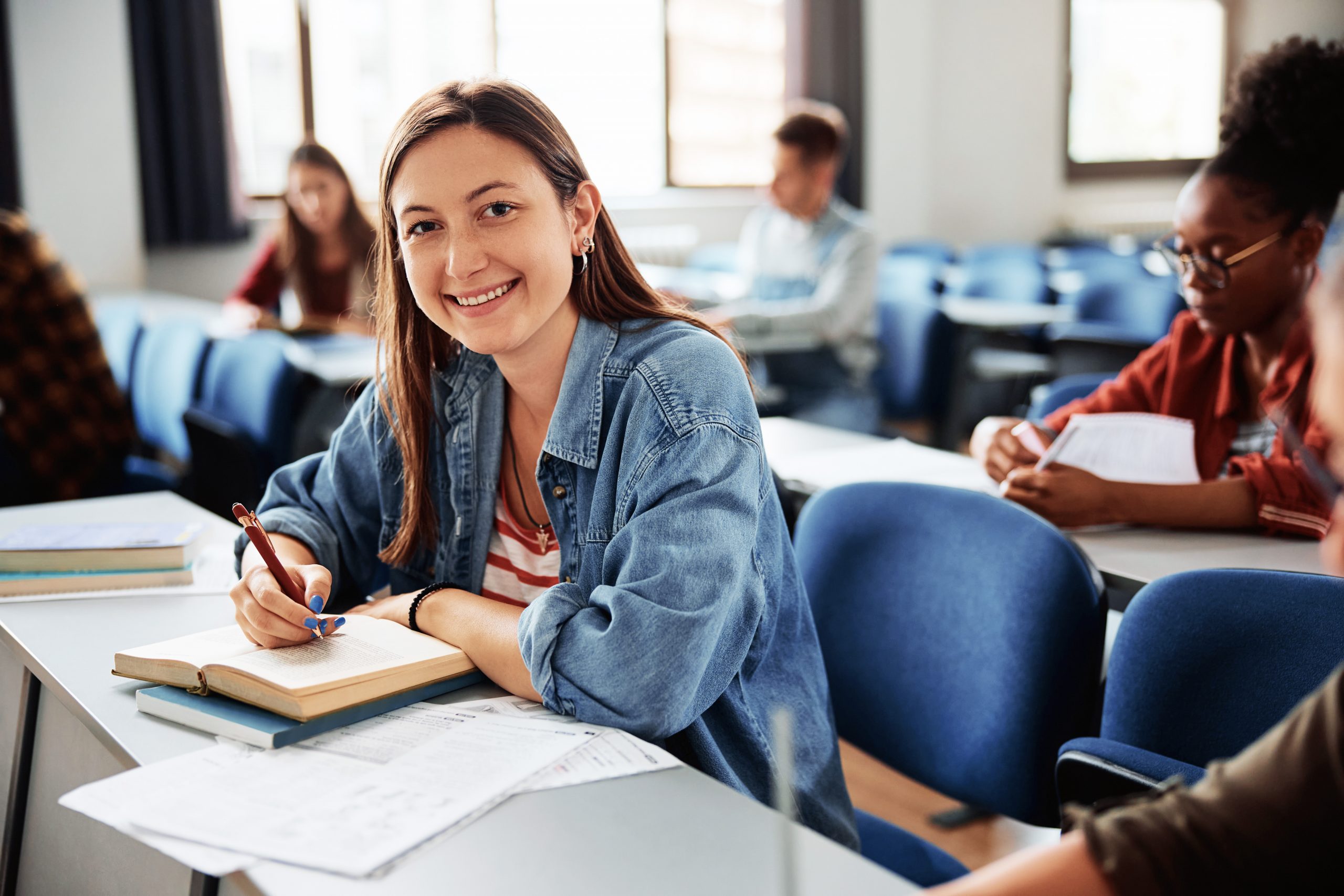Eine Studentin sitzt in einem Seminarraum an einem Tisch, lächelt in die Kamera und schreibt in ein Notizbuch. Im Hintergrund sind weitere Studierende bei der Arbeit zu sehen.