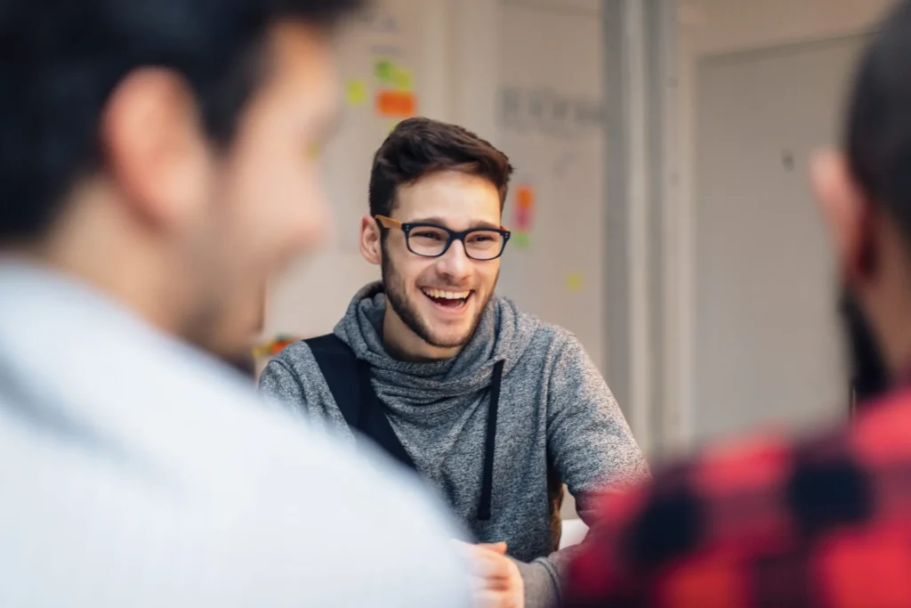 Ein Student mit Brille lacht während eines Gesprächs in einem Seminarraum. Im Vordergrund sind weitere Personen unscharf zu sehen.