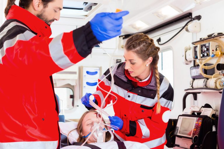 Zwei Rettungskräfte in roter Einsatzkleidung versorgen eine Patientin im Inneren eines Rettungswagens mit medizinischen Geräten.