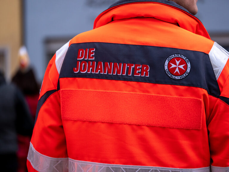 Rückansicht einer Person in einer roten Einsatzjacke mit der Aufschrift „Die Johanniter“ und dem Johanniter-Unfall-Hilfe-Logo.