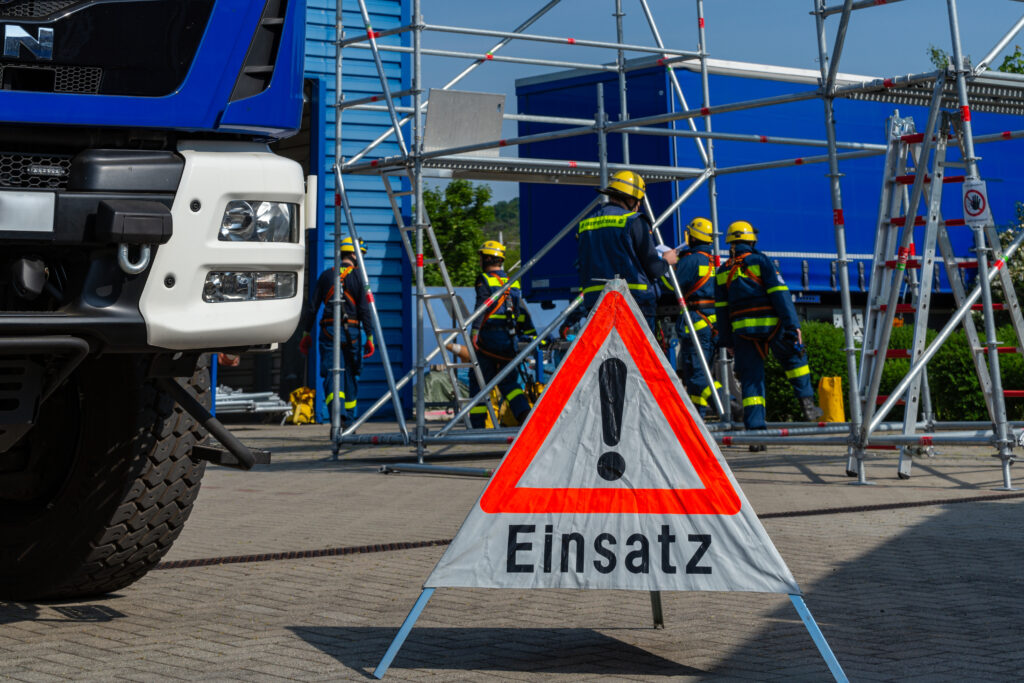 Einsatzkräfte des Technischen Hilfswerks arbeiten an einer Übungsstelle mit Gerüstkonstruktionen. Im Vordergrund steht ein Warnschild mit der Aufschrift „Einsatz“, daneben ein Einsatzfahrzeug.
