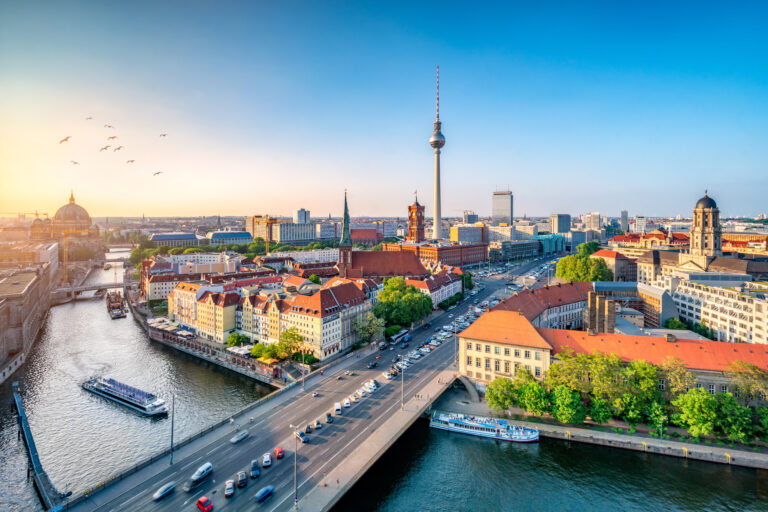 Panoramablick über die Berliner Innenstadt bei Sonnenuntergang mit der Spree im Vordergrund, Schiffen auf dem Fluss, der Museumsinsel links und dem Berliner Fernsehturm sowie dem Roten Rathaus im Zentrum des Bildes.