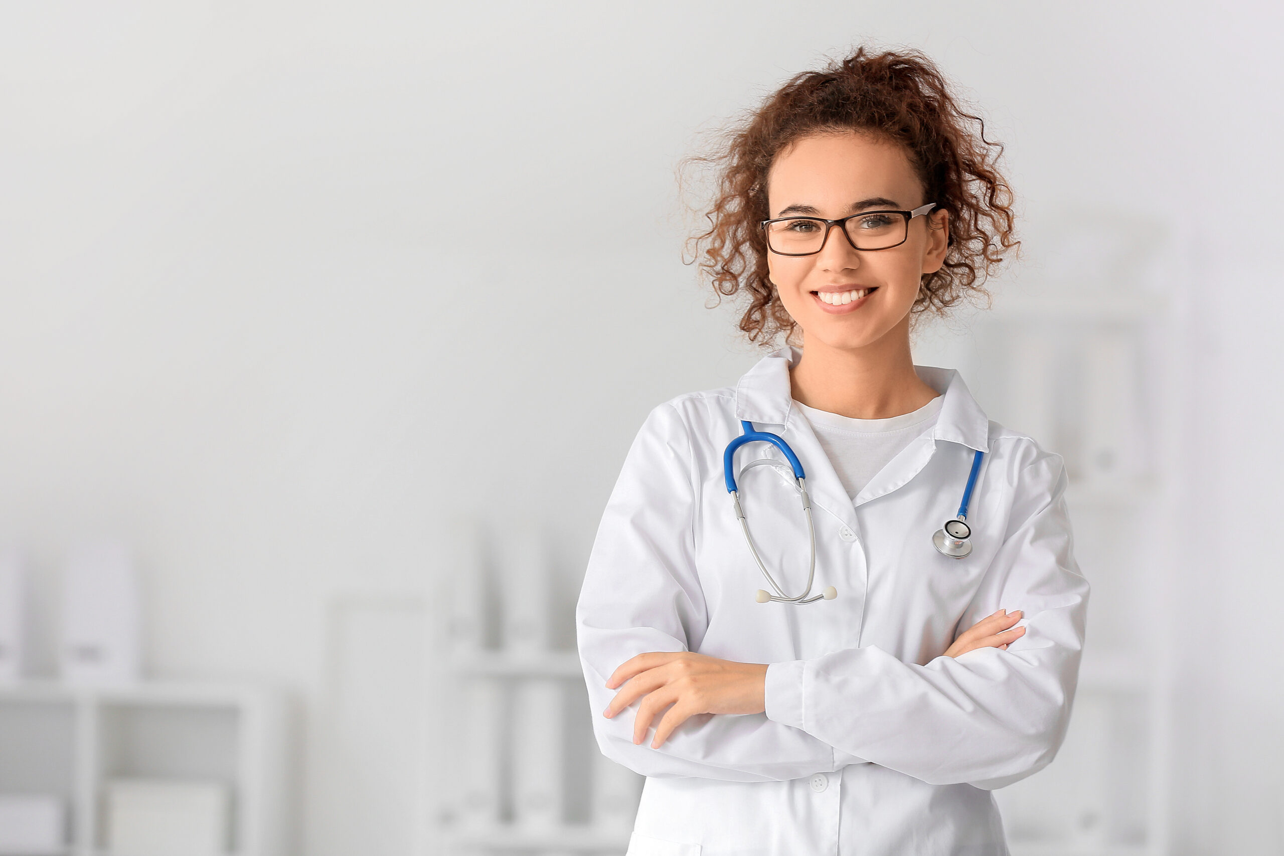 Eine junge Frau in einem weißen Arztkittel steht lächelnd mit verschränkten Armen vor einem unscharfen, hellen Hintergrund eines medizinischen Arbeitsumfelds. Sie trägt eine Brille und hat ein Stethoskop um den Hals.