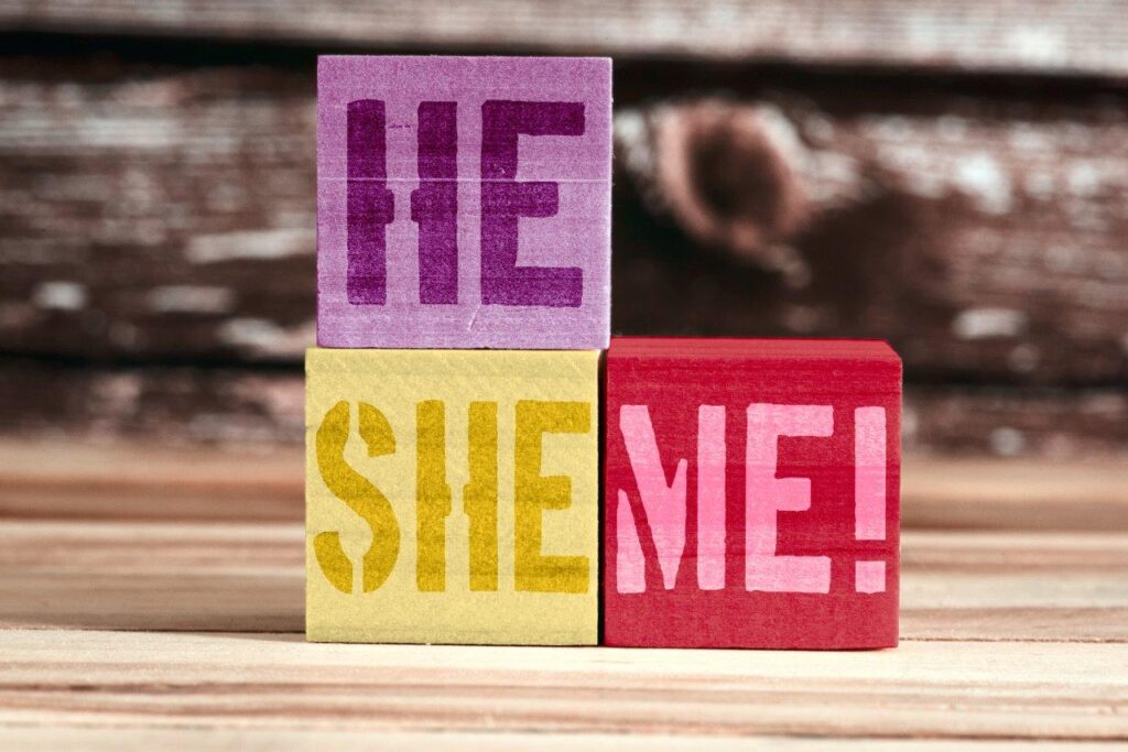 Drei bunte Holzklötze stehen aufeinander bzw. nebeneinander. Auf dem oberen lilafarbenen Block steht „HE“, darunter auf einem gelben Block „SHE“ und daneben auf einem roten Block „ME!“. Die Klötze liegen auf einer Holzoberfläche, im Hintergrund ist eine unscharfe Holzwand zu sehen.