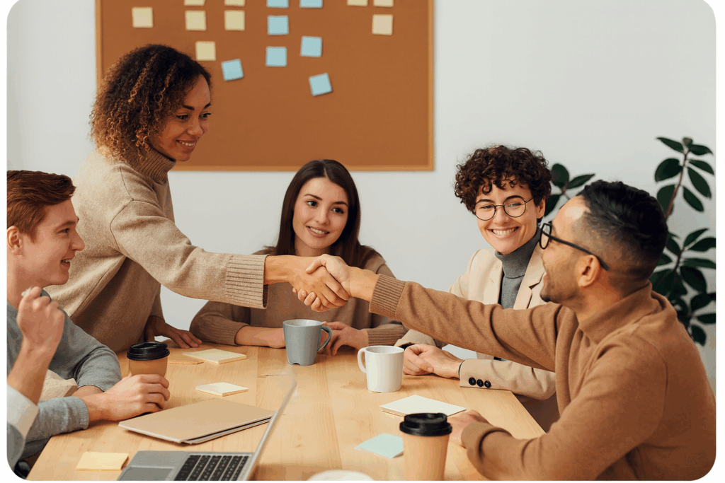 Mehrere junge Erwachsene sitzen an einem Tisch in einem Besprechungsraum und geben sich über den Tisch hinweg die Hand. Sie wirken freundlich und engagiert; auf dem Tisch liegen Notizzettel, Tassen und ein Laptop.