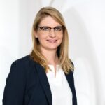 Porträtfoto einer lächelnden Frau mit blonden, schulterlangen Haaren und Brille. Sie trägt ein dunkelblaues Sakko über einer weißen Bluse. Der Hintergrund ist hell und unscharf, sodass die Person im Vordergrund klar hervortritt.