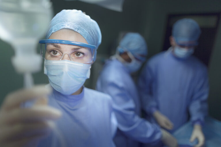 Eine Person in vollständiger OP-Schutzkleidung – inklusive Haube, Maske und Schutzbrille – fokussiert auf eine Infusion, die sie vorbereitet oder überprüft. Im Hintergrund arbeiten zwei weitere Personen in blauer OP-Kleidung an einem Operationstisch. Die Szene spielt in einem beleuchteten Operationssaal.