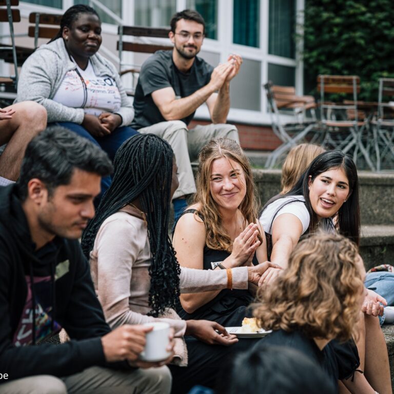 Studierende sitzen in einer gemischten Gruppe auf Treppenstufen im Außenbereich des Campus und tauschen sich in entspannter Atmosphäre aus. Einige halten Tassen oder Teller in der Hand, im Vordergrund steht der persönliche Austausch und das gemeinschaftliche Lernen.