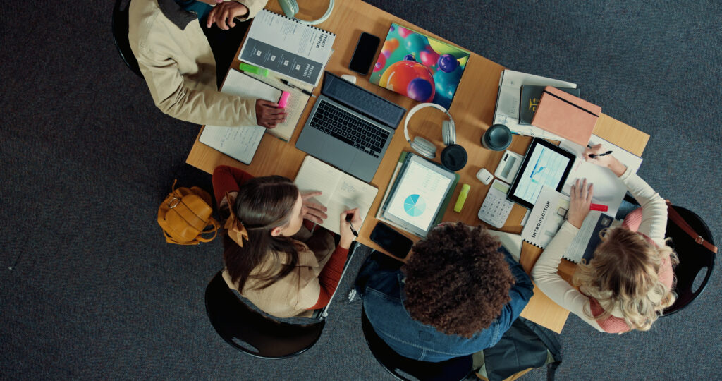 Vier Studierende sitzen an einem Tisch und arbeiten gemeinsam an Laptops, Tablets und Notizbüchern. Auf dem Tisch liegen Lernmaterialien, Diagramme, Kopfhörer und Schreibutensilien, die auf eine kooperative Lern- und Arbeitssituation im Hochschulkontext hinweisen.