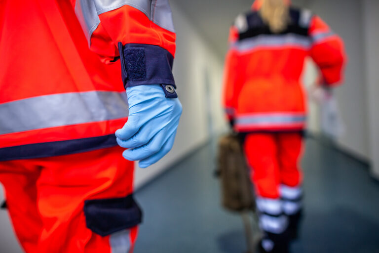 Rettungsdienstpersonal in Schutzkleidung auf einem Flur, Fokus auf eine behandschuhte Hand im Vordergrund.
