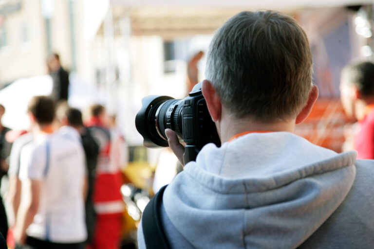 Eine Person fotografiert mit einer professionellen Kamera eine Veranstaltung im öffentlichen Raum. Im Hintergrund sind mehrere Menschen unscharf zu sehen, darunter Einsatzkräfte in roter Schutzkleidung, was auf eine Übungs- oder Einsatzsituation im Bereich Bevölkerungsschutz oder Katastrophenhilfe hinweist.